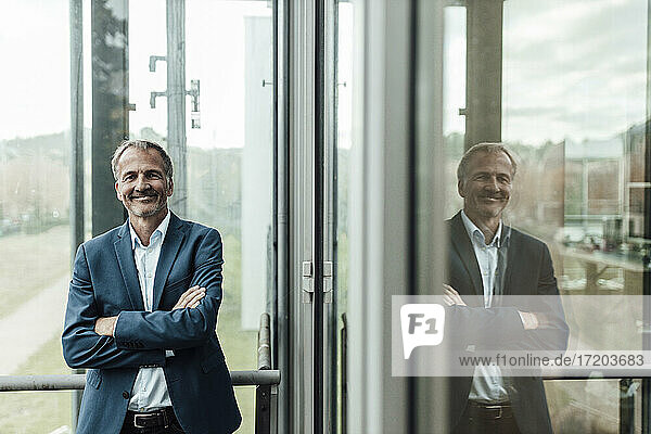 Lächelnder männlicher Geschäftsmann mit verschränkten Armen vor einem Fenster im Büro stehend