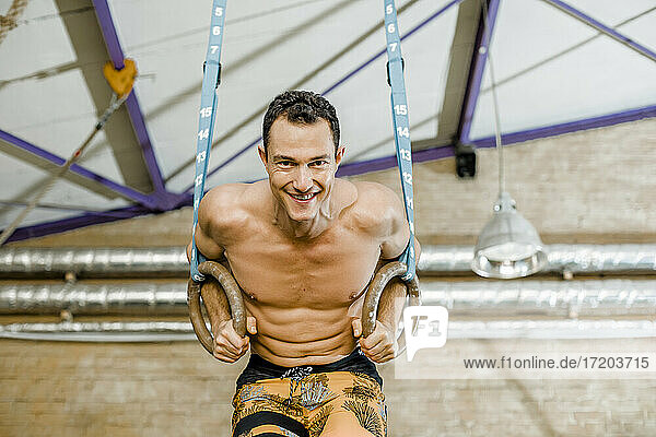 Lächelnder Mann hängt mit Gymnastikringen an der Wand im Fitnessstudio