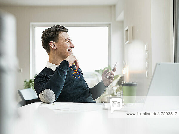 Lächelnder männlicher Unternehmer  der im Büro zu Hause sitzt und über sein Smartphone im Internet surft