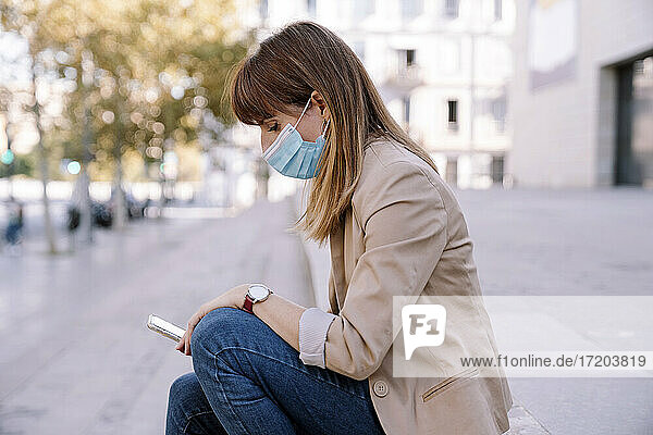 Junge Frau mit Gesichtsschutzmaske  die im Freien sitzend ihr Smartphone benutzt