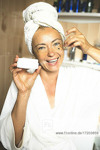 Lächelnde reife Frau trägt im Badezimmer Feuchtigkeitscreme auf das Gesicht auf