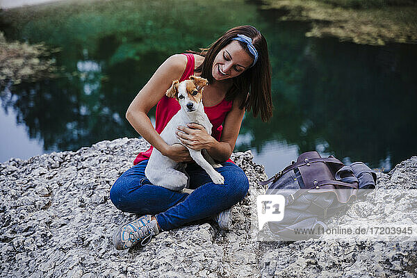Verspielte Frau hält ihren Hund und sitzt auf einem Felsen am See