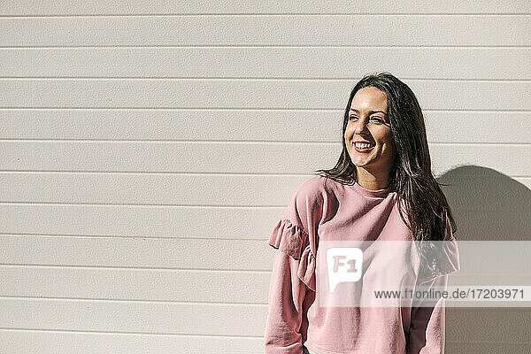 Smiling woman looking away while standing against wall during sunny day