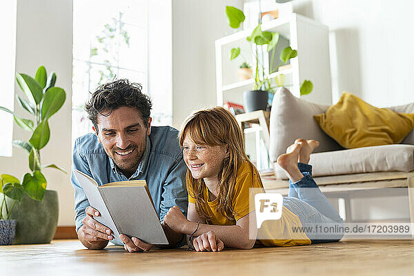 Smiling father and daughter reading book together while lying down on floor at home