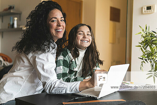 Smiling mother and daughter with laptop sitting at table in living room