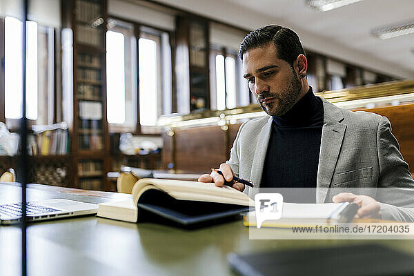 Geschäftsmann mit Laptop  der ein Buch liest  während er in einer Bibliothek arbeitet