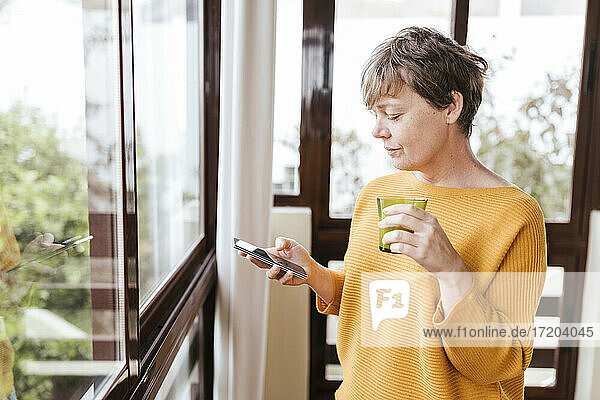 Reife Frau mit Trinkglas  die ihr Smartphone benutzt  während sie am Glasfenster steht