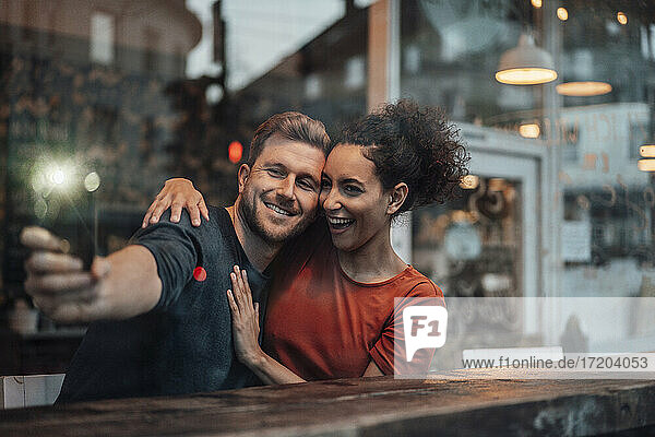Fröhliches Paar nimmt Selfie durch Handy beim Sitzen im Café