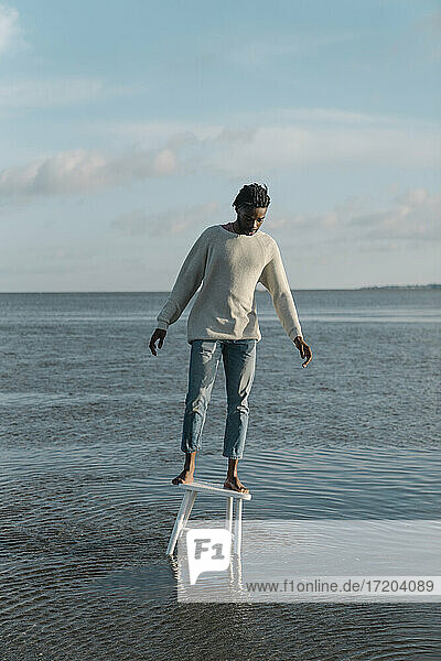 Junger Mann steht auf einem weißen Hocker und balanciert am Strand gegen den bewölkten Himmel