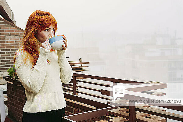 Junge rothaarige Frau trinkt Kaffee auf der Terrasse und schaut weg