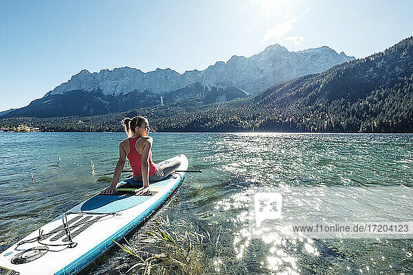 Deutschland  Bayern  Garmisch Partenkirchen  Junge Frau sitzt auf einem Stand Up Paddle Board auf dem Eibsee und schaut auf die Zugspitze