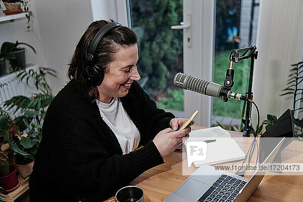 Lächelnde Frau  die während einer Podcast-Sitzung zu Hause ihr Smartphone neben dem Laptop am Schreibtisch benutzt