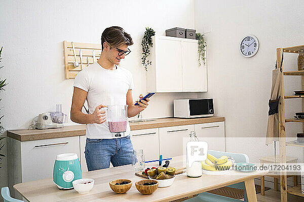 Junger Mann hält einen Smoothie-Mixer in der Hand und benutzt sein Smartphone in der Küche