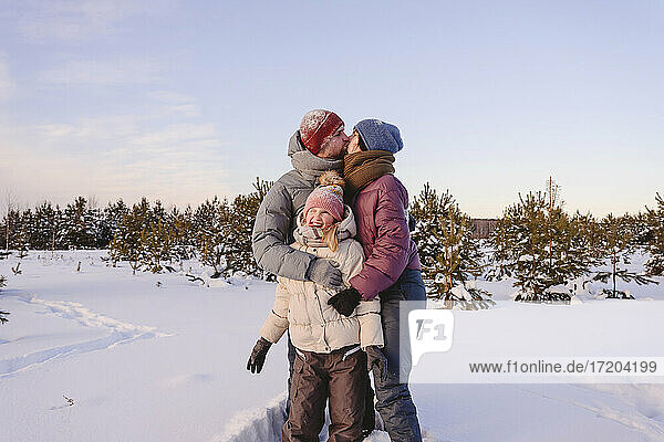 Küssende Eltern  die ihre süße Tochter im Winter im Schnee umarmen