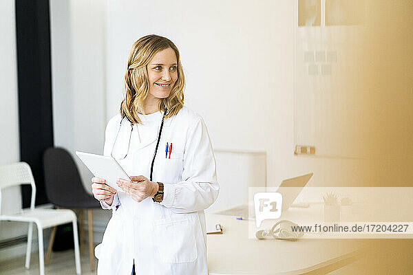 Lächelnde Mitarbeiterin des Gesundheitswesens mit Tablet  die sich in einer medizinischen Klinik auf den Schreibtisch stützt