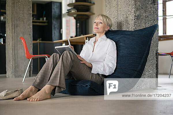 Businesswoman with digital tablet looking away while relaxing on floor chair at home
