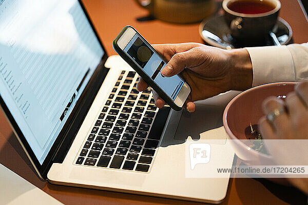 Geschäftsmann  der ein Smartphone über einem Laptop benutzt  während er in einem Café sitzt