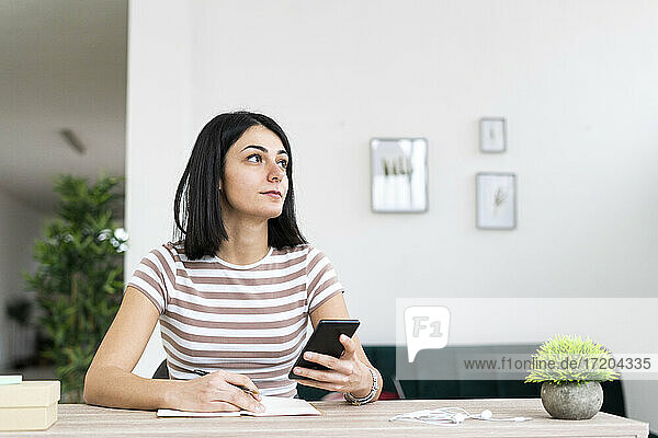 Nachdenkliche junge Frau mit Smartphone sitzt am Tisch im Wohnzimmer