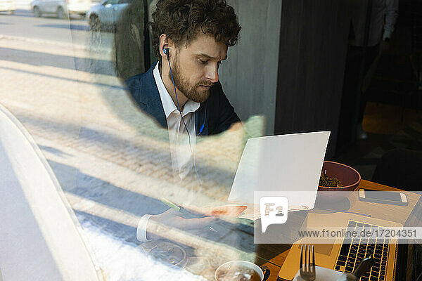 Männlicher Unternehmer  der während eines Videogesprächs am Laptop im Café sitzt und eine Strategie liest
