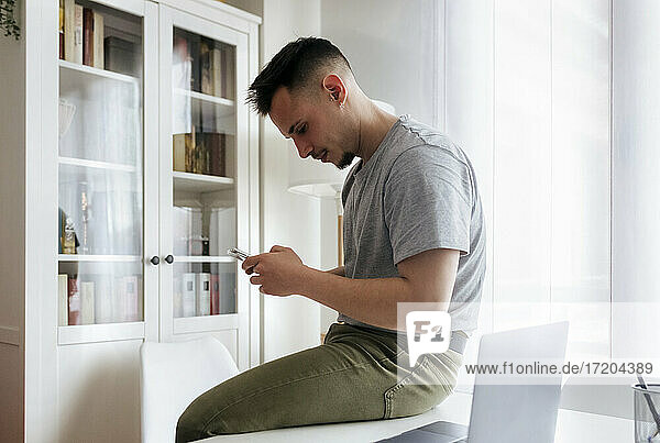 Gut aussehender Mann  der ein Smartphone benutzt  während er im Büro zu Hause auf dem Schreibtisch sitzt