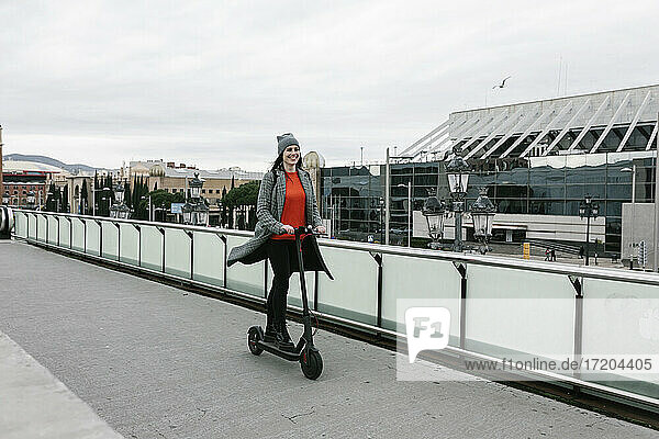 Glückliche Frau fährt elektrischen Roller auf dem Gehweg in der Nähe des Geländers in der Stadt