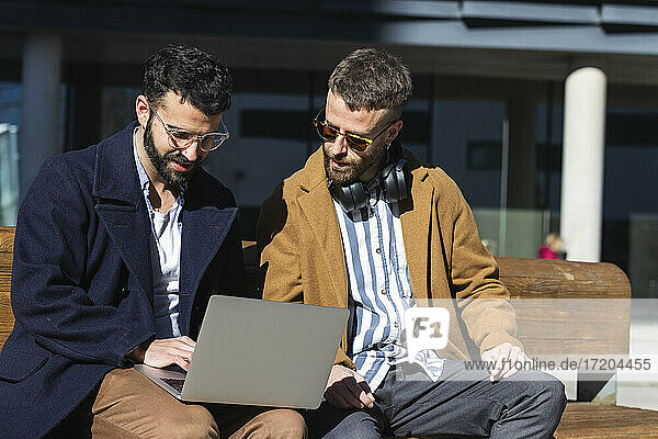 Geschäftspartner arbeiten an einem Laptop  während sie an einem sonnigen Tag auf einer Bank sitzen