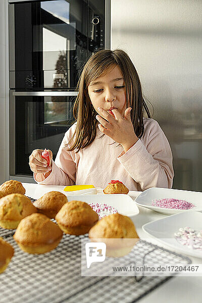 Niedliches Mädchen leckt Finger beim Dekorieren von Muffins am Küchentisch