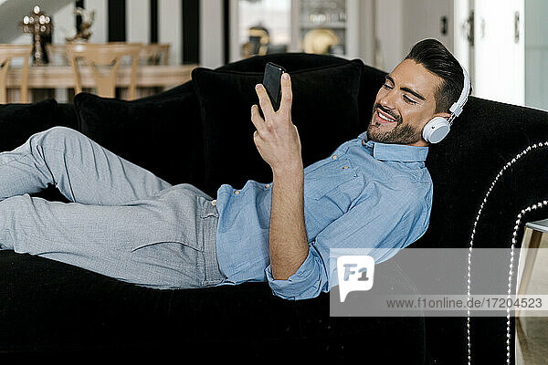 Lächelnder Mann mit Kopfhörern  der ein Mobiltelefon benutzt  während er sich zu Hause auf dem Sofa ausruht