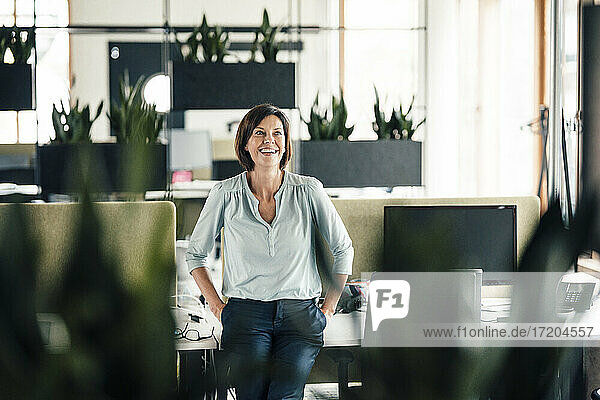 Lächelnde Geschäftsfrau mit Händen in den Taschen  die sich im Büro auf den Schreibtisch stützt