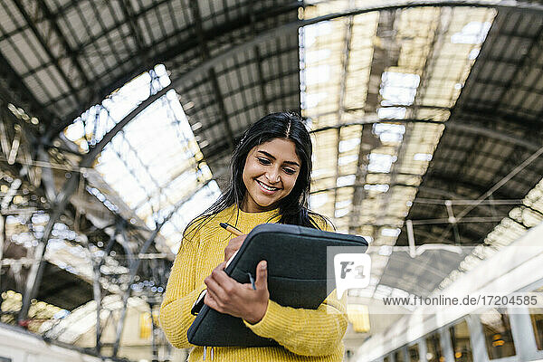 Lächelnde Frau mit Laptoptasche  die auf dem Bahnsteig in ihr Tagebuch schreibt