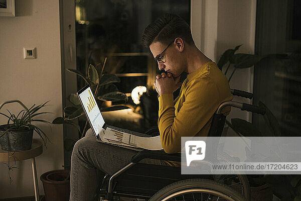 Concentrated oung man in wheelchair with laptop working late while sitting on wheelchair in living room