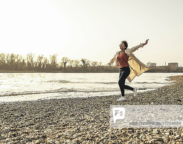 Cheerful woman wearing overcoat running by lake on sunset