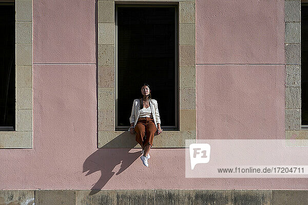 Junge Frau sitzt an einem sonnigen Tag auf der Fensterbank eines Gebäudes