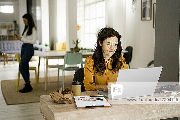 Reife Geschäftsfrau,  die an einem Laptop arbeitet,  während ihre Tochter im Büro zu Hause ein Mobiltelefon benutzt