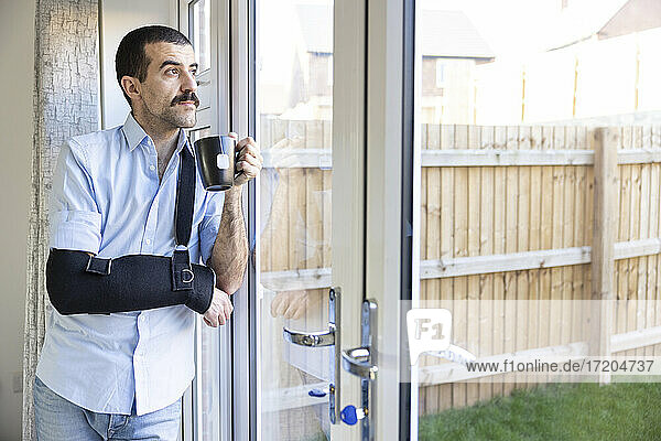 Thoughtful man wearing arm sling drinking tea while looking through window at home