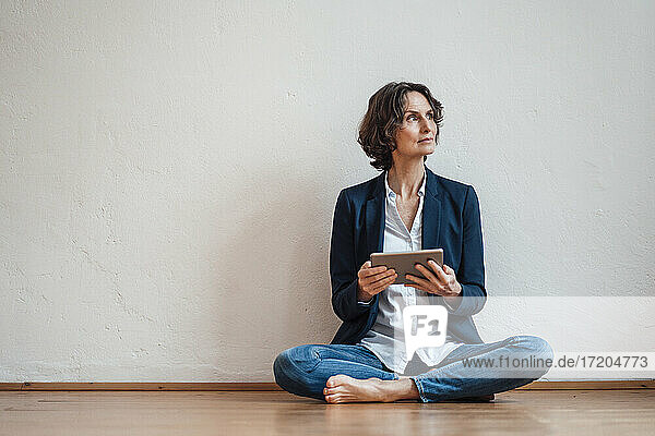 Nachdenkliche Geschäftsfrau mit digitalem Tablet auf dem Boden vor einer weißen Wand sitzend