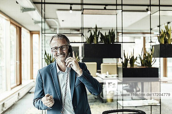 Smiling businessman with eyeglasses talking on telephone while standing in office