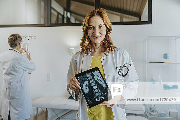 Arzt zeigt Röntgenbild auf digitalem Tablet  während er mit einem Mitarbeiter im Hintergrund in einer Klinik steht