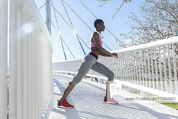 Junge Sportlerin  die sich die Beine vertritt  während sie auf einer Brücke an einem sonnigen Tag übt