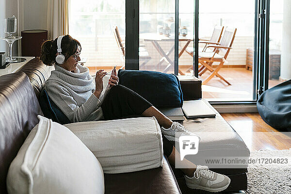 Reife Frau mit Kopfhörern  die ein Mobiltelefon benutzt  während sie zu Hause sitzt