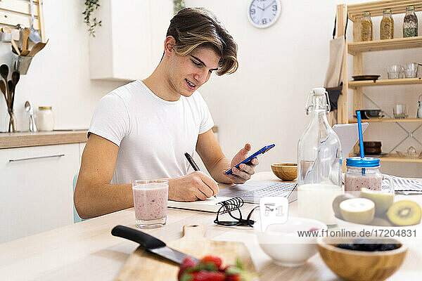 Young man with smart phone writing on notepad while sitting at table in kitchen