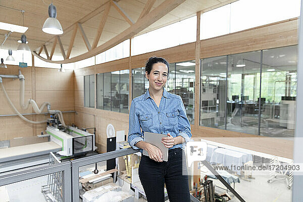Smiling businesswoman with digital tablet leaning on railing in factory