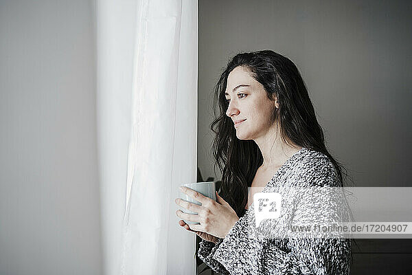 Nachdenklich lächelnde Frau  die eine Kaffeetasse hält  während sie durch ein Fenster zu Hause schaut
