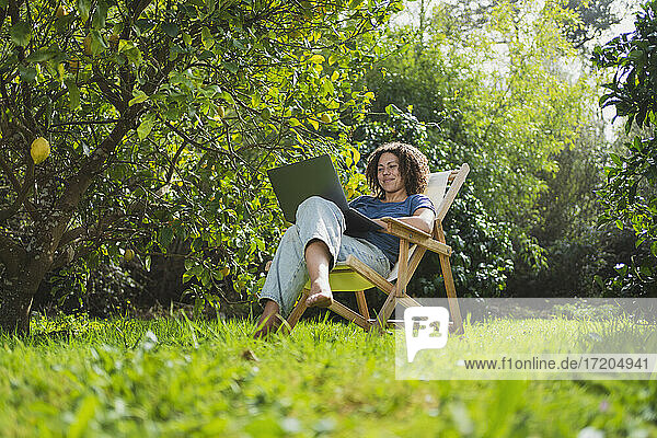 Lächelnde Frau schaut auf einen Laptop  während sie auf einem Stuhl im Permakultur-Garten sitzt