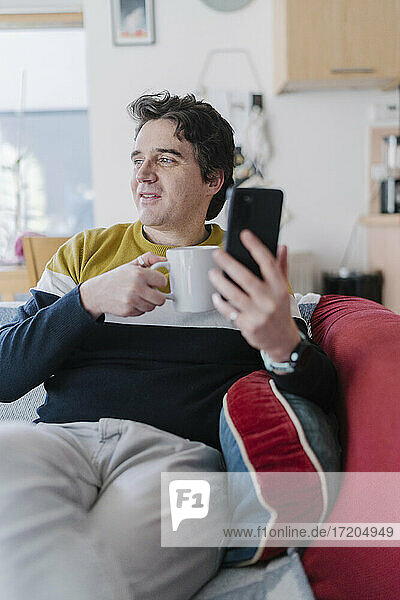 Mann mit Smartphone und Kaffeetasse auf dem Sofa sitzend und wegschauend