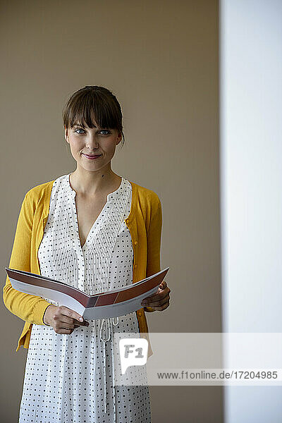 Lächelnde Geschäftsfrau mit Dokumenten an der Wand stehend im Büro