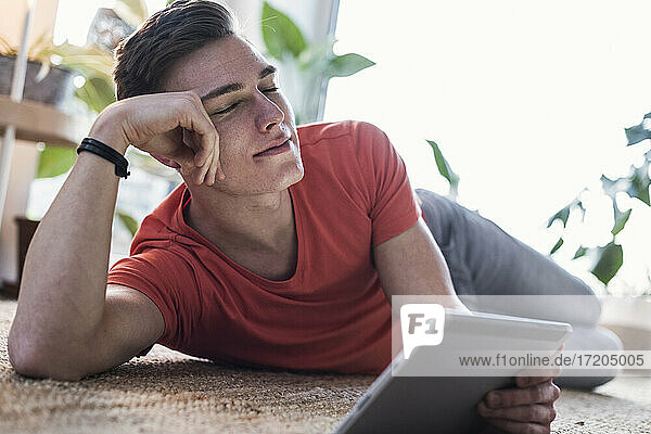 Entspannter junger Mann mit digitalem Tablet auf dem Teppich im Wohnzimmer liegend