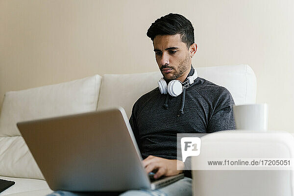 Junger Mann mit Kopfhörern  der einen Laptop benutzt  während er zu Hause auf dem Sofa sitzt