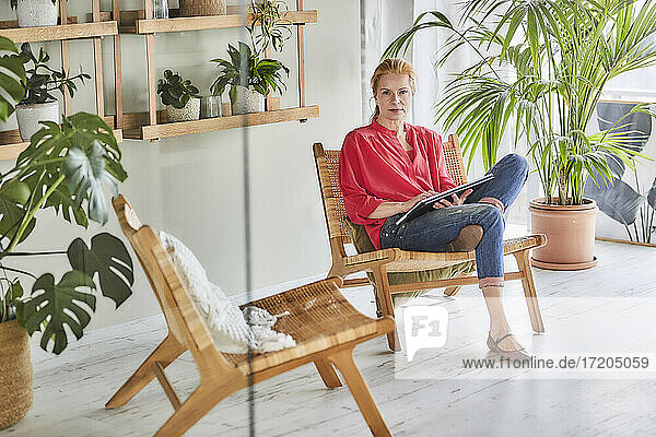 Mature woman in casual clothing with laptop sitting on chair in loft apartment at home