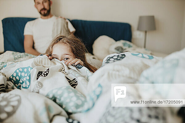 Mädchen versteckt sich unter einer Decke  während der Vater zu Hause auf dem Bett zusieht